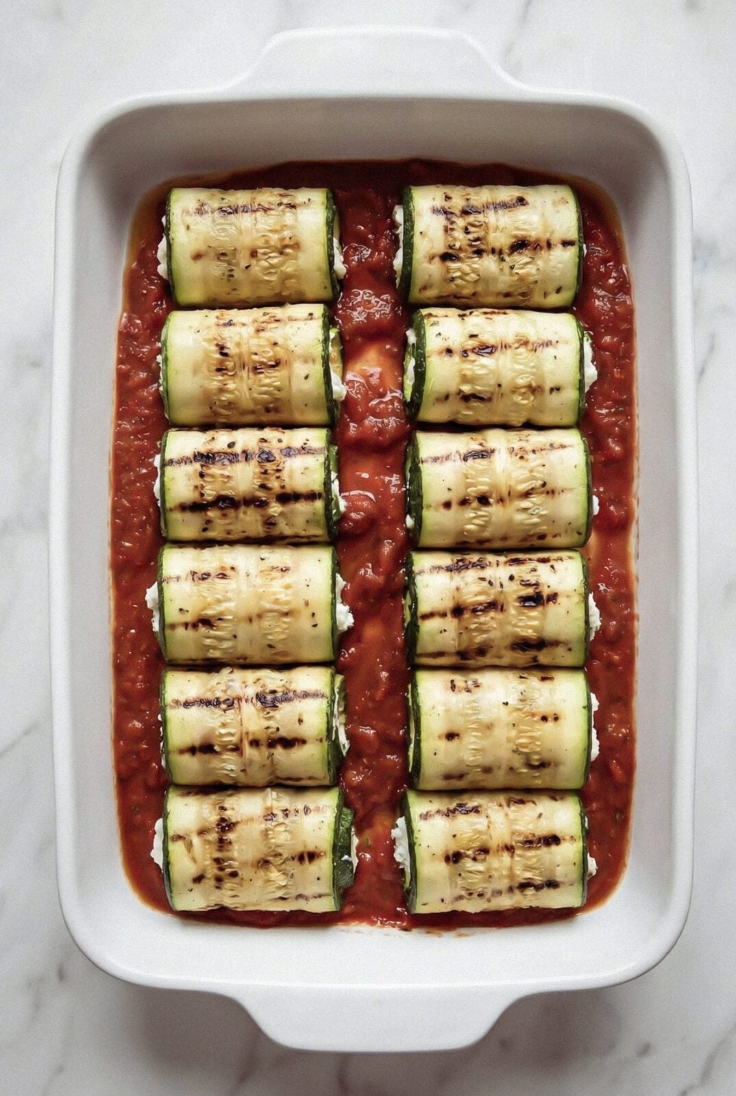 Zucchini rolls arranged seam side down over marinara in a baking dish