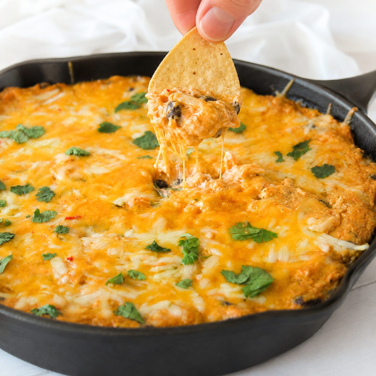 A hand dips a tortilla chip into a skillet of chicken enchilada dip