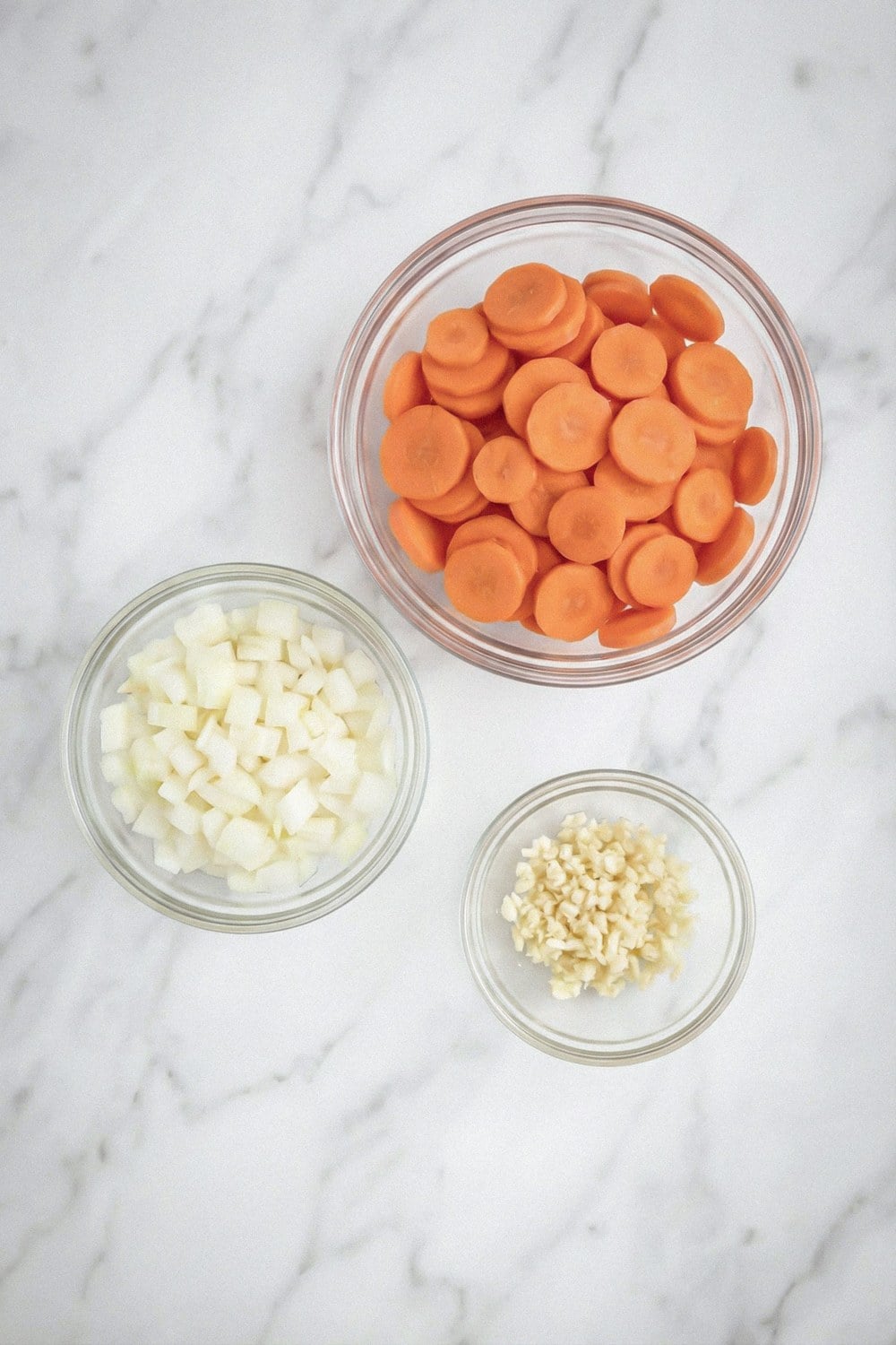 Sliced carrots, diced onion, and minced garlic in glass bowls