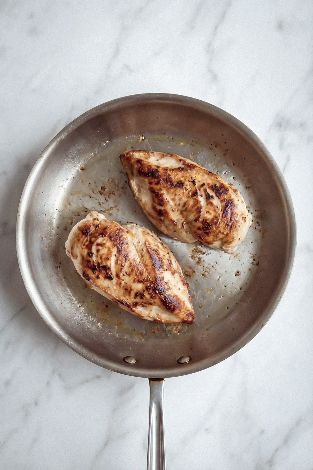 Two seared chicken breasts in a stainless steel skillet