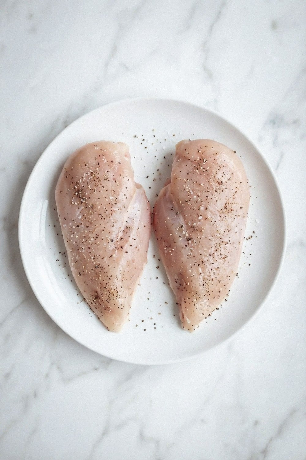 Two raw chicken breasts seasoned with salt and pepper on a plate
