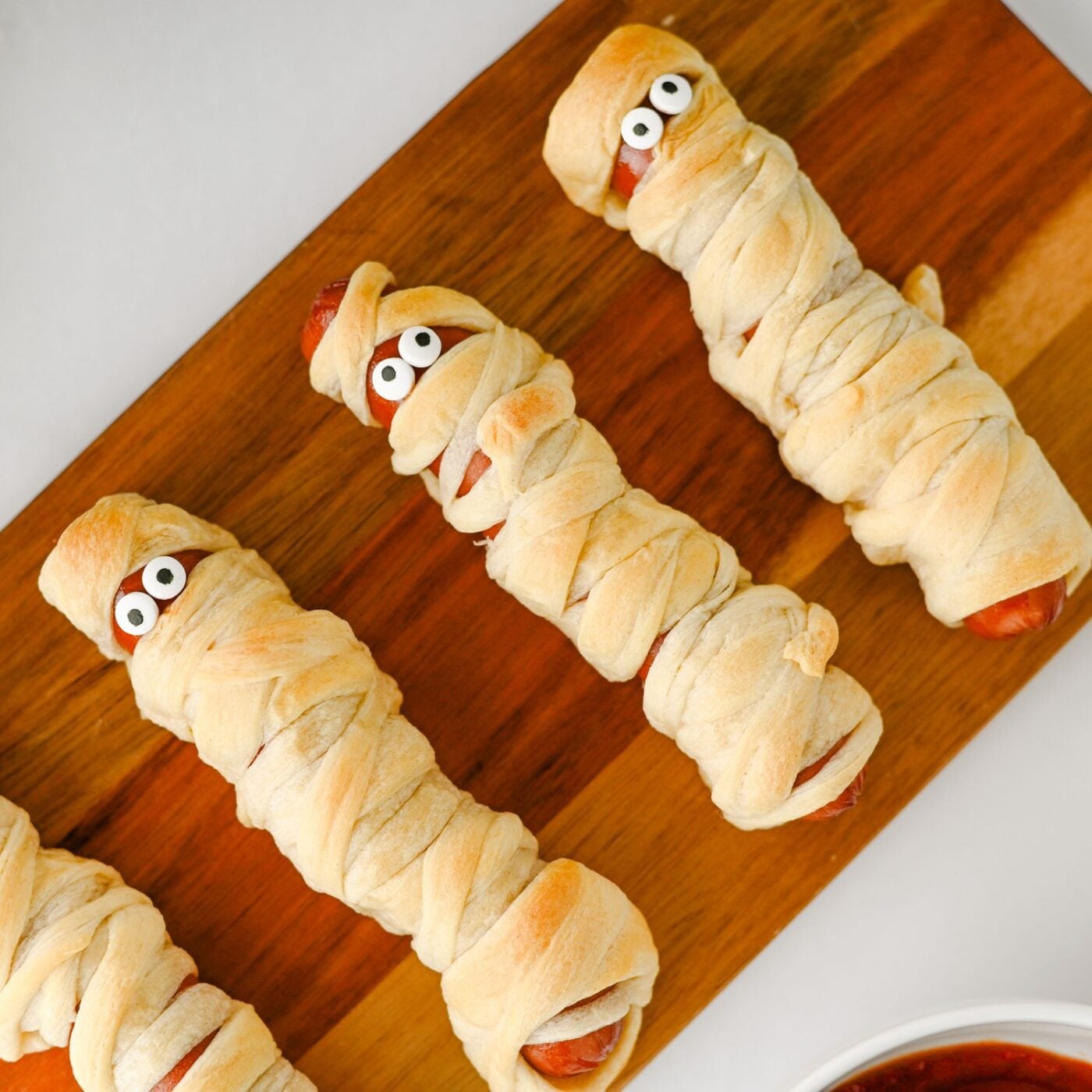 Mummy hot dogs wrapped in crescent roll dough with candy eyes on a wooden board