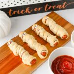 Mummy hot dogs on a wooden board with a trick or treat sign and ketchup