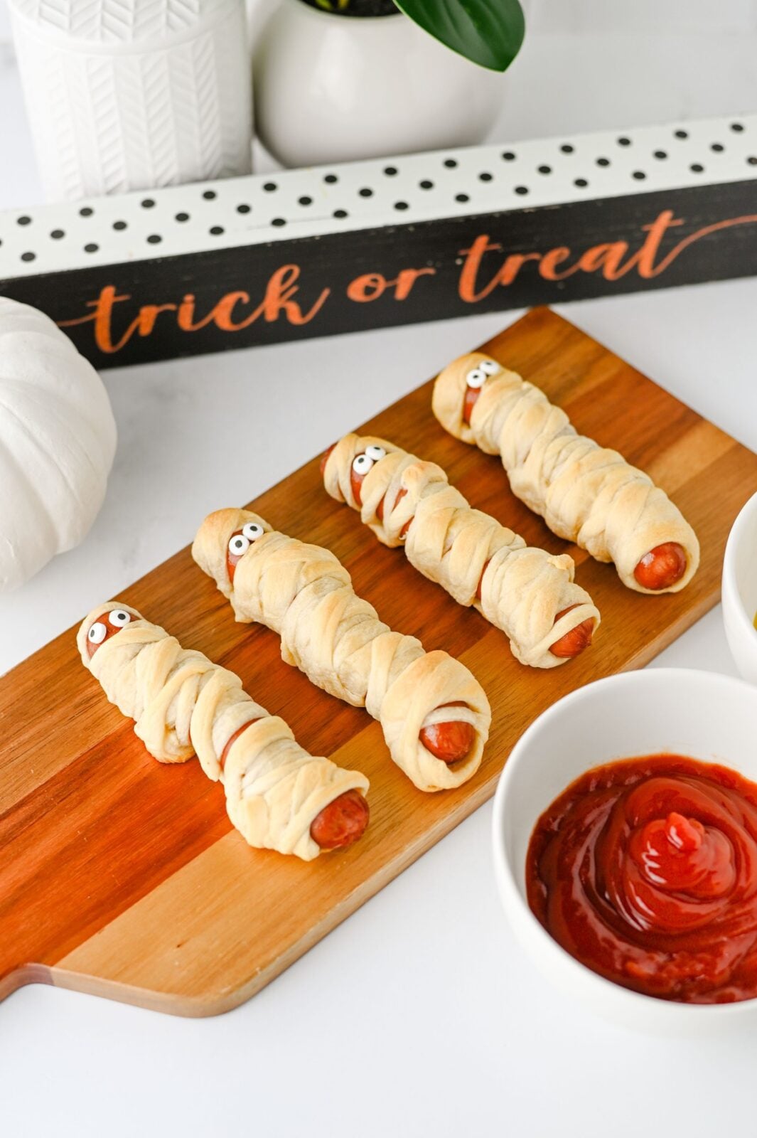 Mummy hot dogs on a wooden board with a trick or treat sign and ketchup