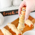 Hand holding a mummy hot dog above the serving board
