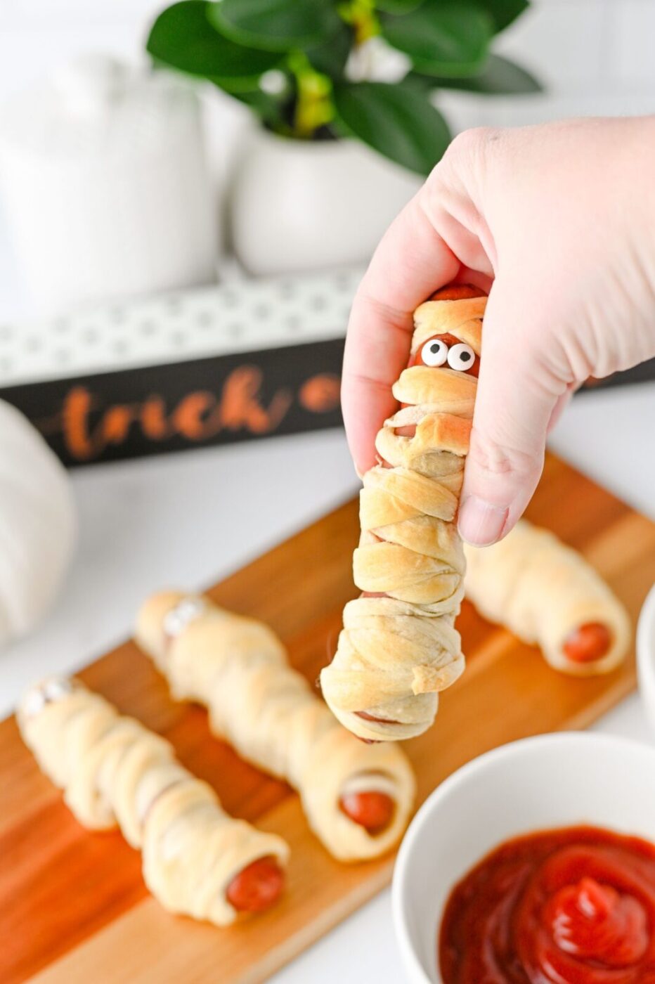 Hand holding a mummy hot dog above the serving board