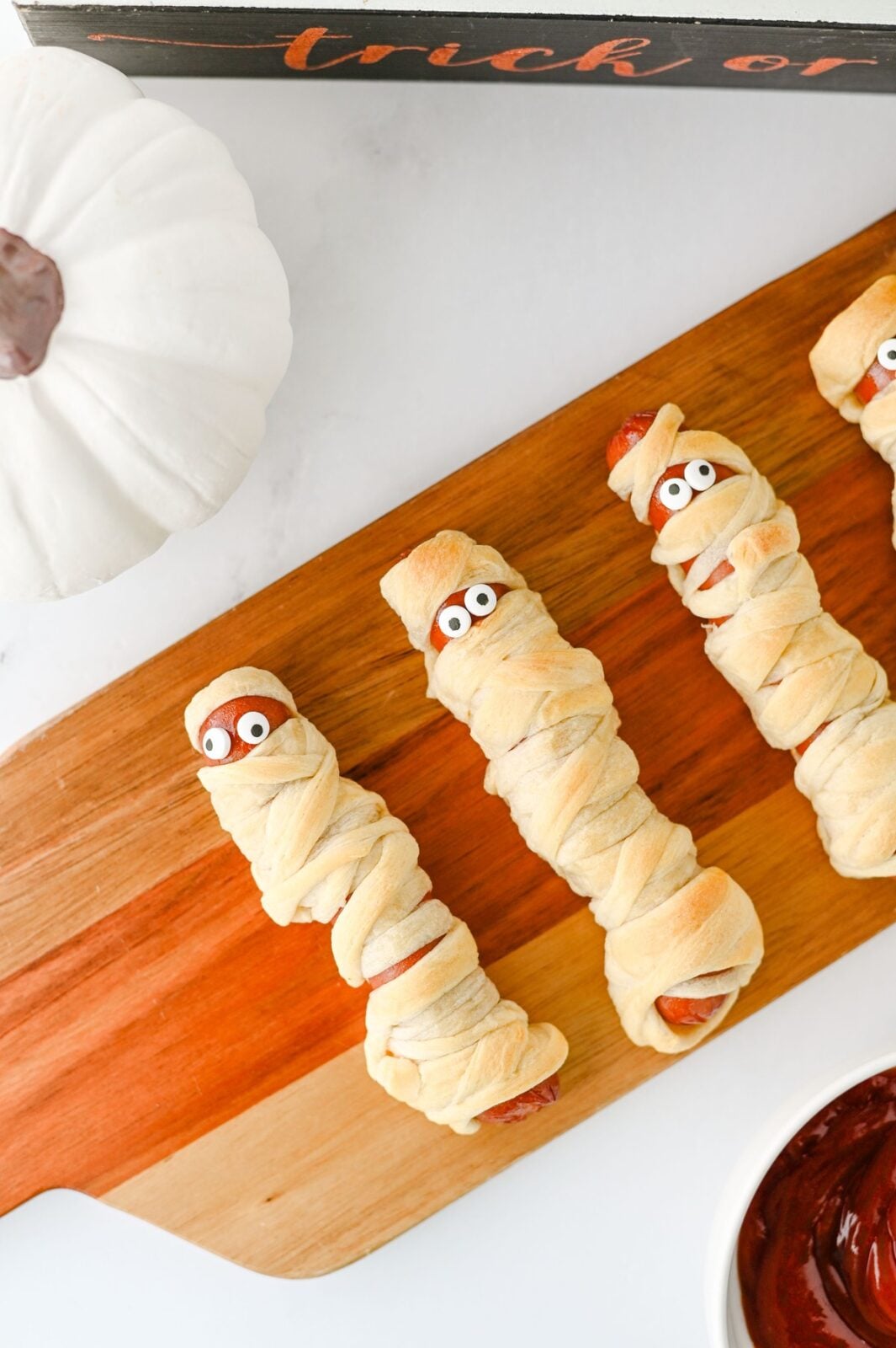 Four mummy hot dogs on a serving board with a white pumpkin