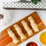 Mummy hot dogs with candy eyes on a wooden board with ketchup and mustard for dipping