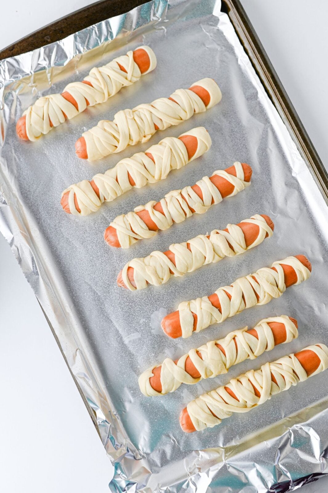 Unbaked mummy hot dogs lined up on a foil-lined baking sheet
