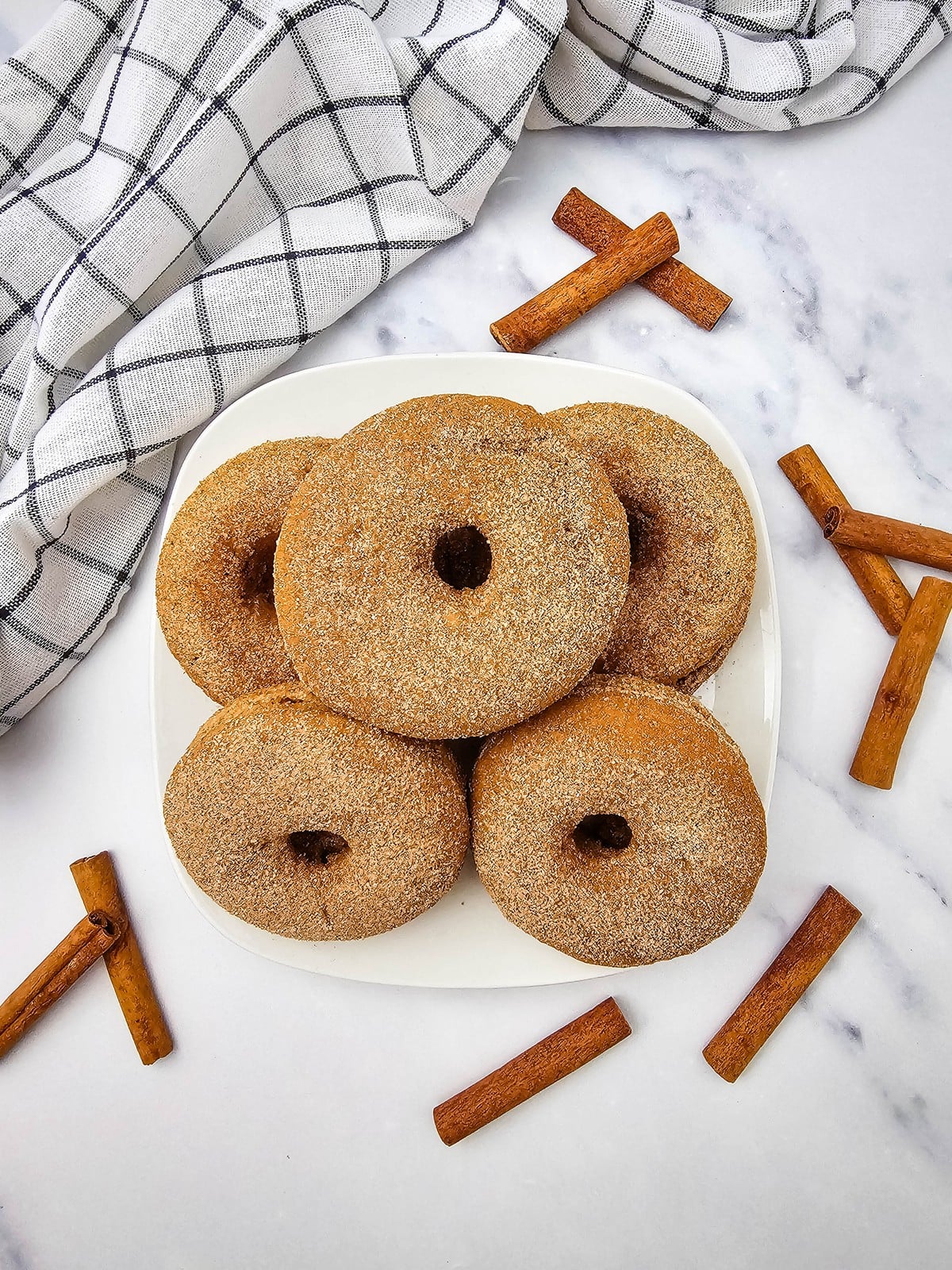 Baked apple cider donuts coated in cinnamon sugar on a white plate with cinnamon sticks
