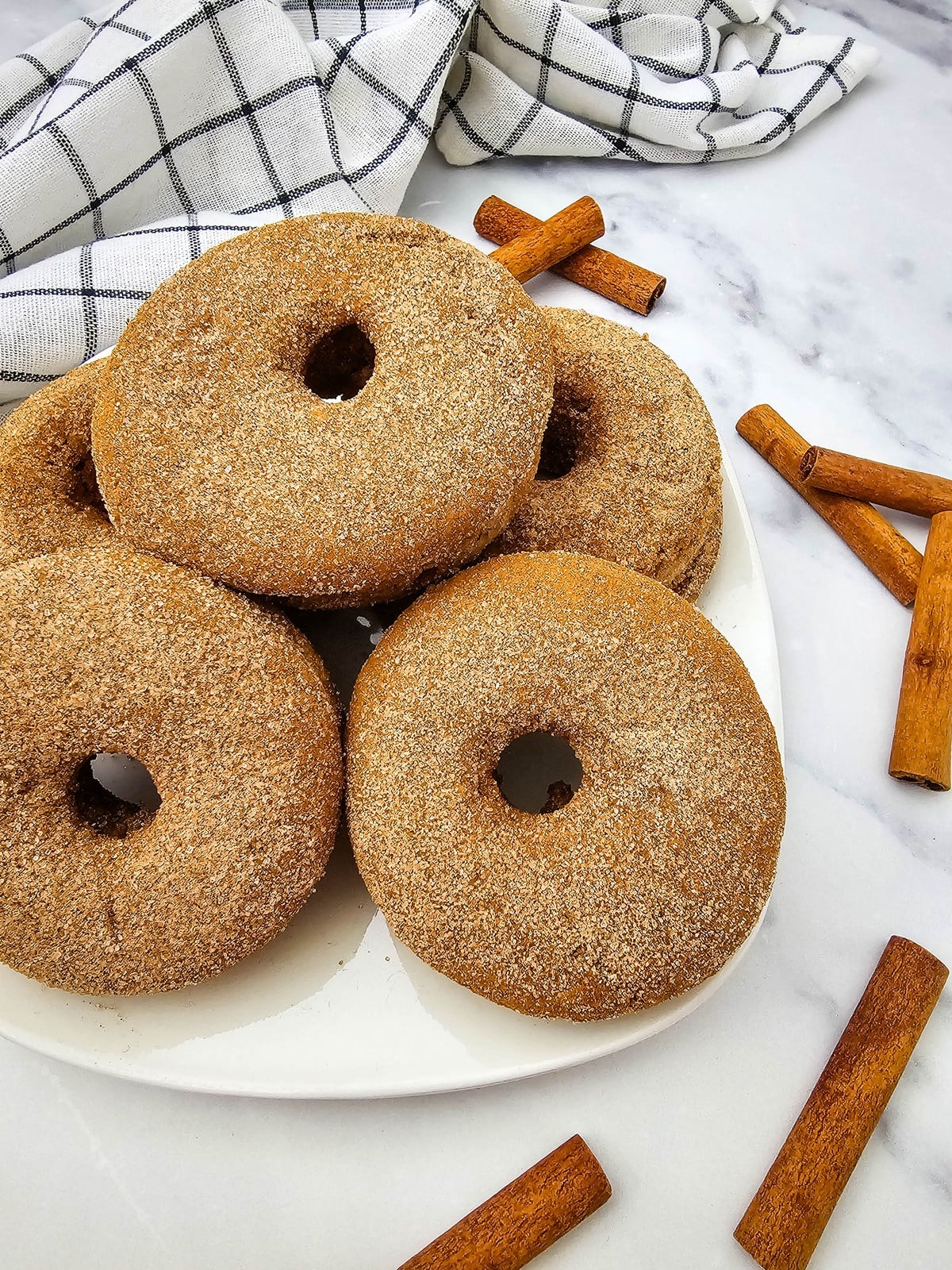 Stack of baked apple cider donuts on a white plate with a checked towel and cinnamon sticks