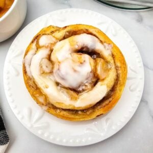 Single apple cinnamon roll on a white plate with icing over the spiral