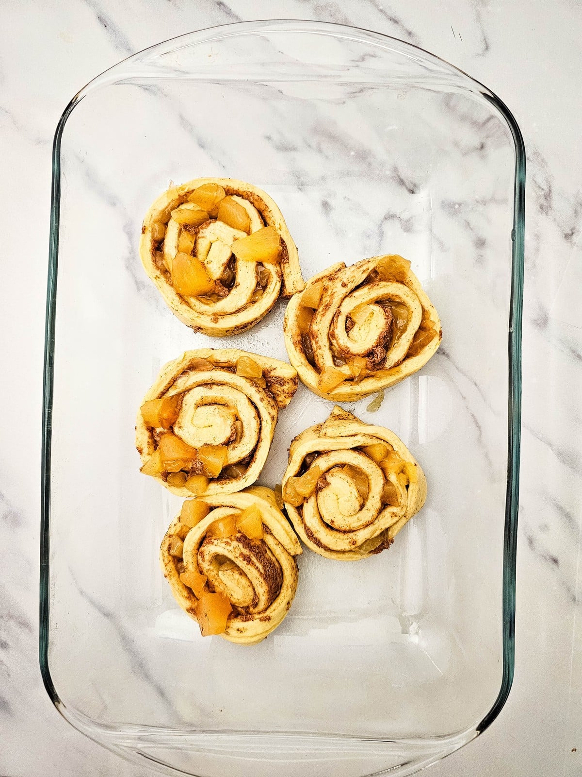 Five unbaked apple cinnamon rolls arranged in a glass baking dish