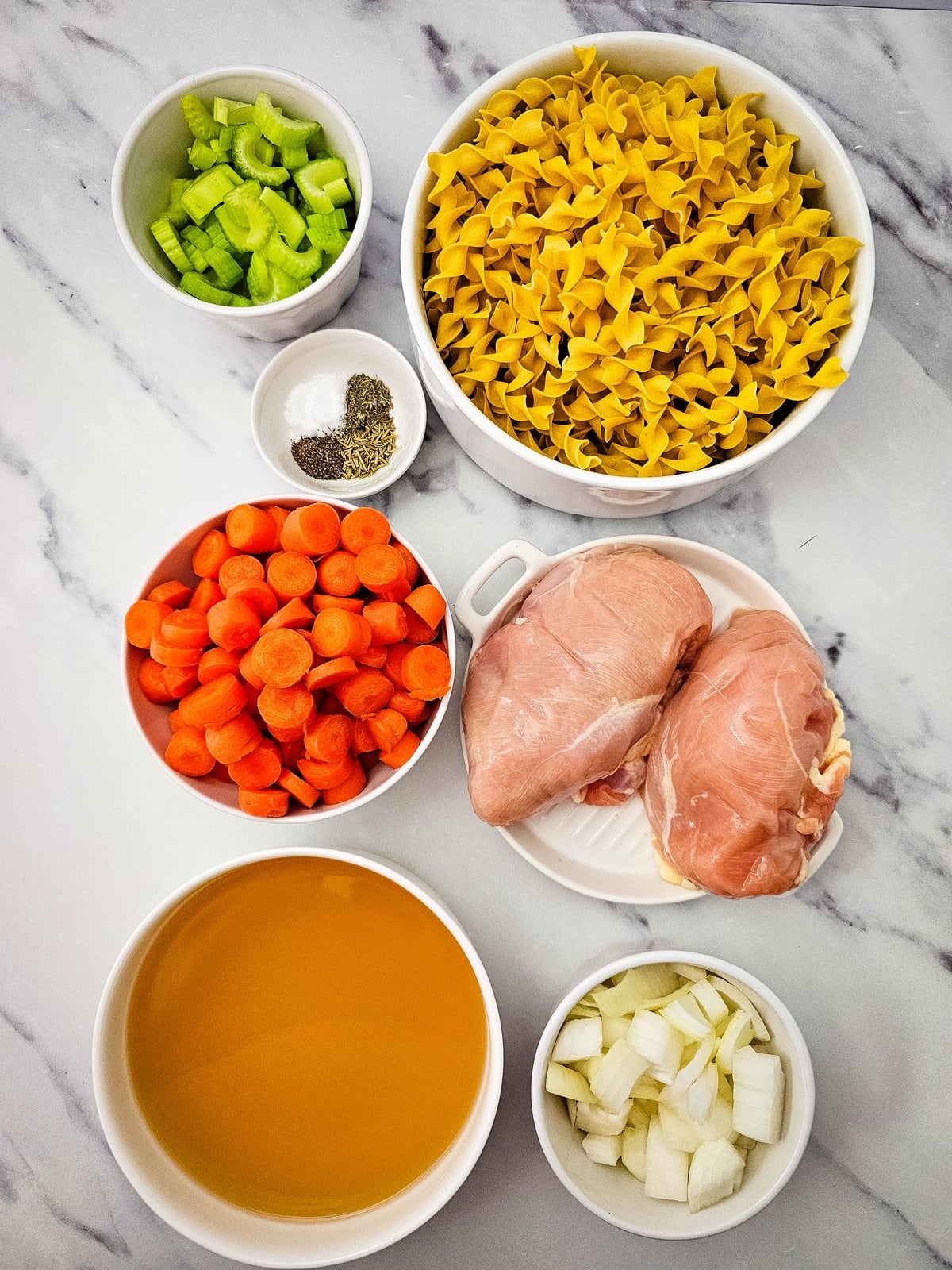 everything you need laid out: chicken breasts, sliced carrots, diced celery, diced onion, chicken broth, seasonings and wide egg noodles