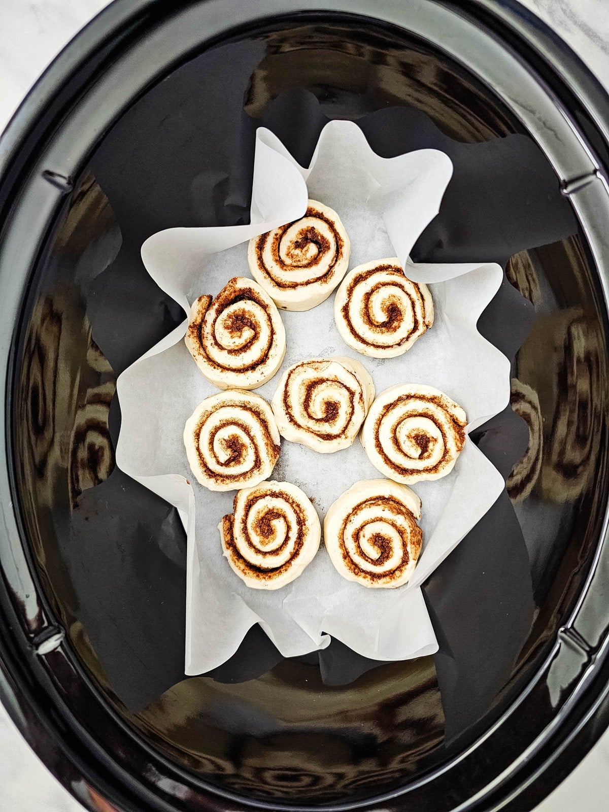 Raw cinnamon rolls arranged on parchment paper in a black slow cooker insert