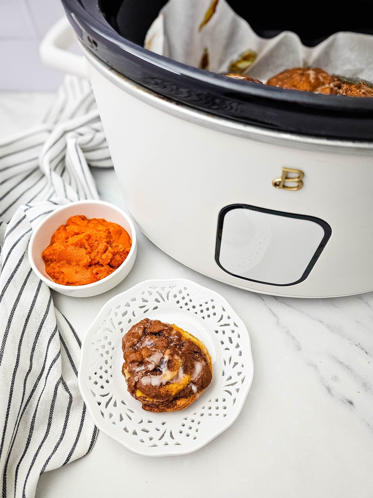 A single crockpot pumpkin cinnamon roll on a white plate beside the slow cooker and a bowl of pumpkin puree