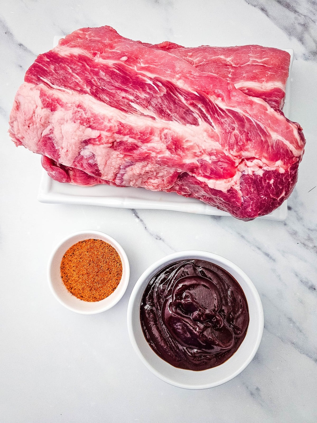 Baby back ribs, BBQ seasoning, and barbecue sauce laid out on a marble counter