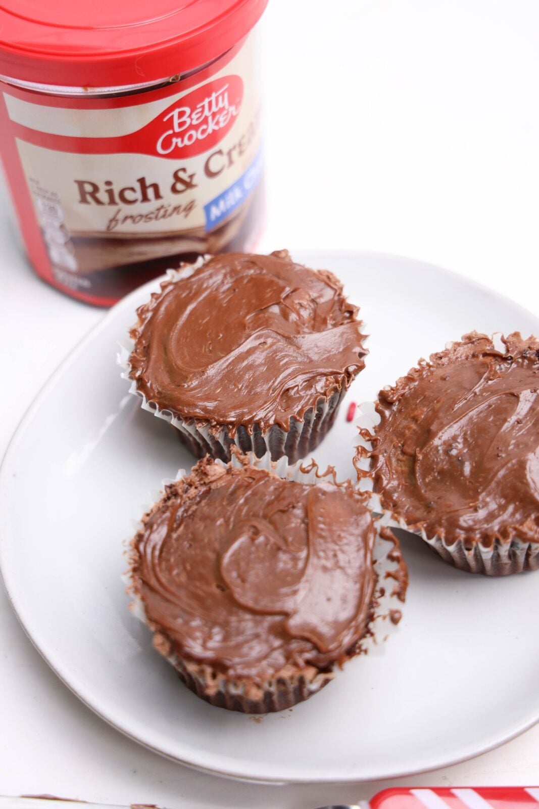 Three frosted chocolate cupcakes on a white plate in front of a tub of chocolate frosting
