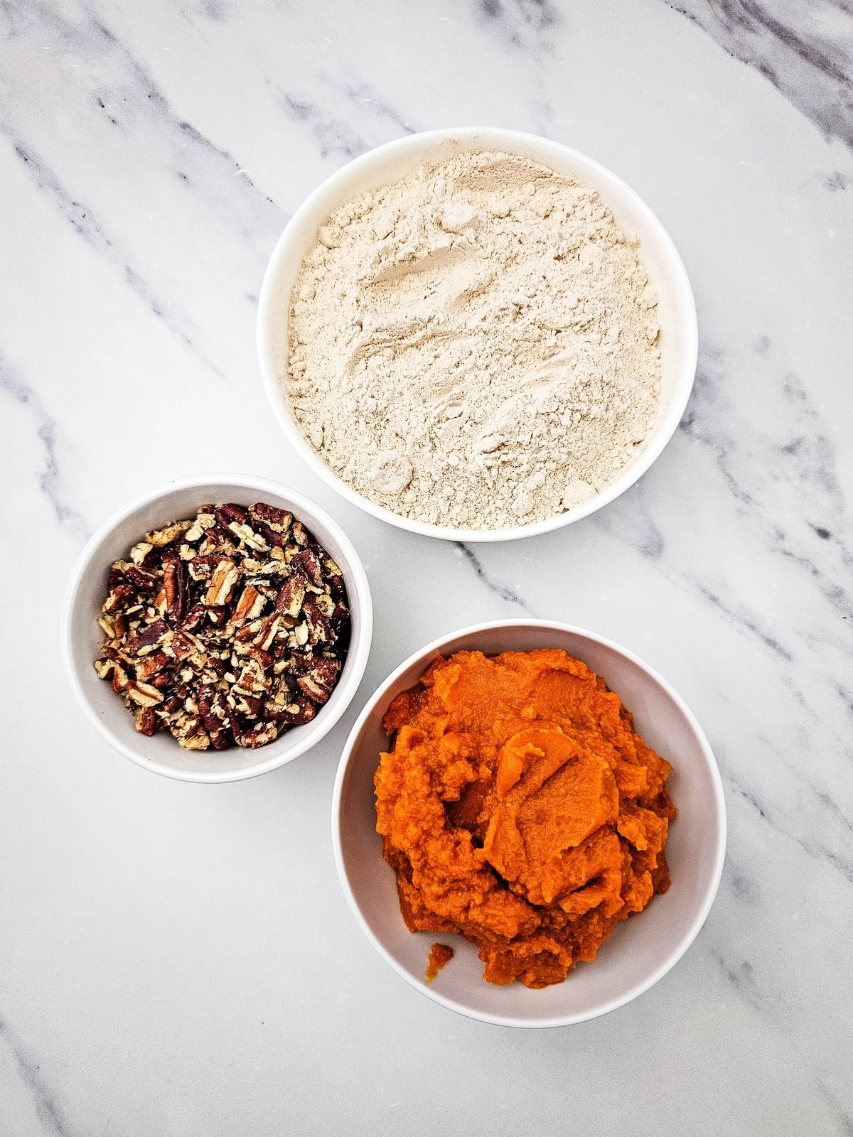 Spice cake mix, chopped pecans, and canned pumpkin puree in three white bowls on marble