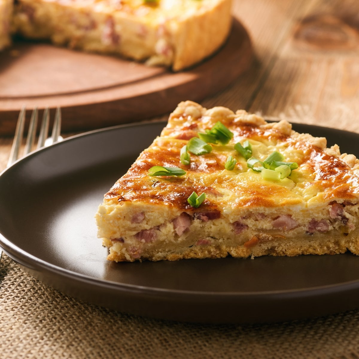 Slice of ham and cheese quiche on a dark plate with green onions, ready to serve with sides
