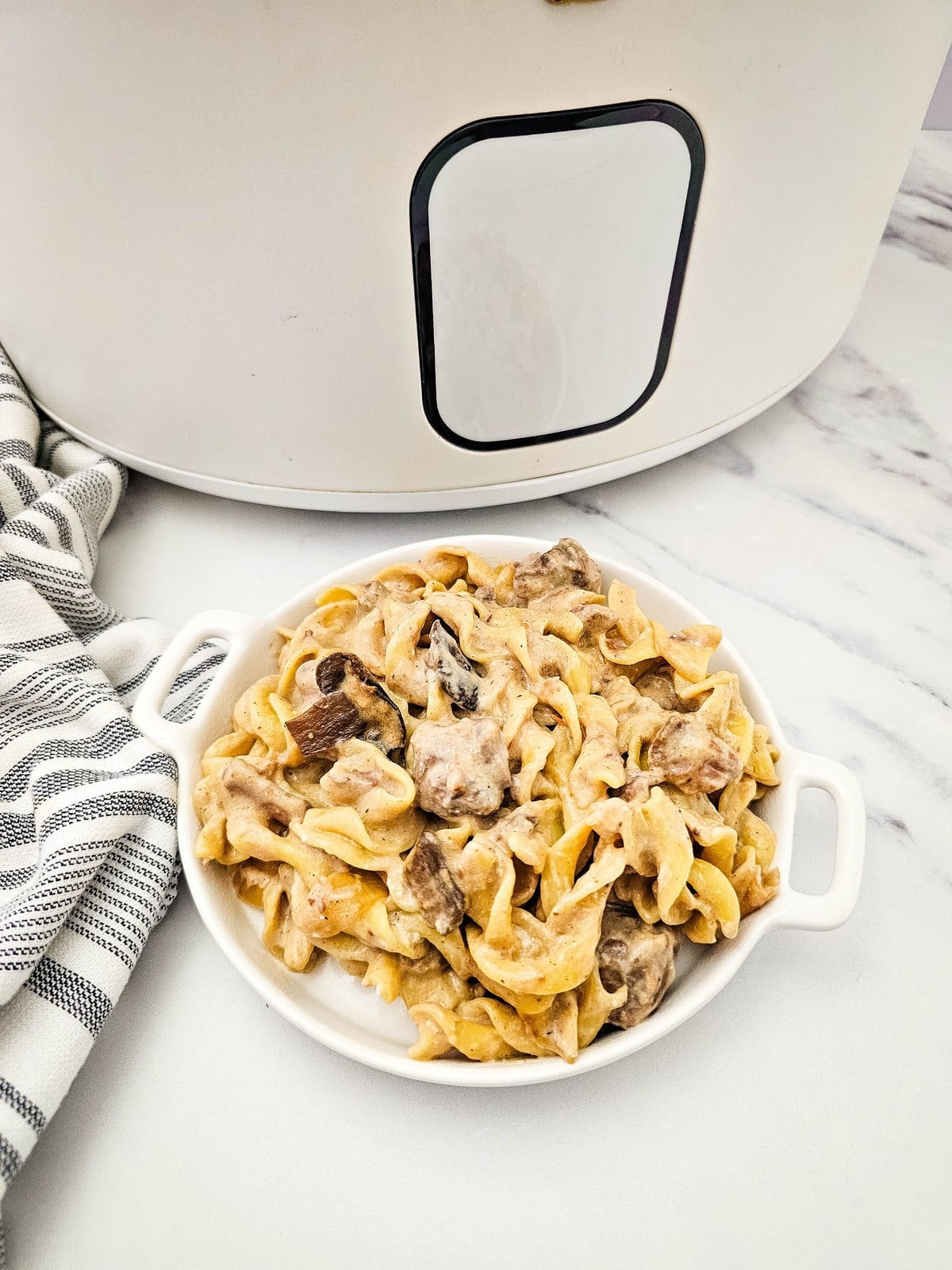 A white handled dish of beef stroganoff on a marble counter beside a white slow cooker and a striped towel