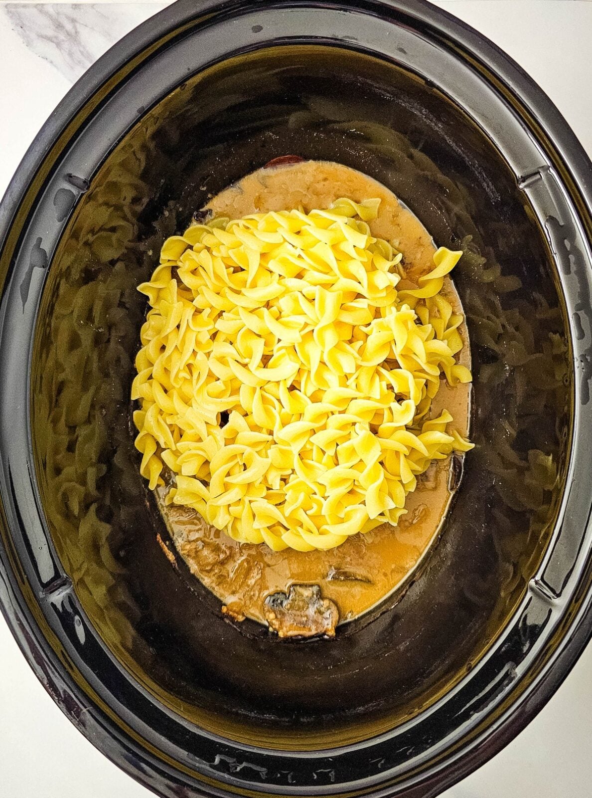 Drained cooked egg noodles piled on top of the finished stroganoff sauce in the slow cooker