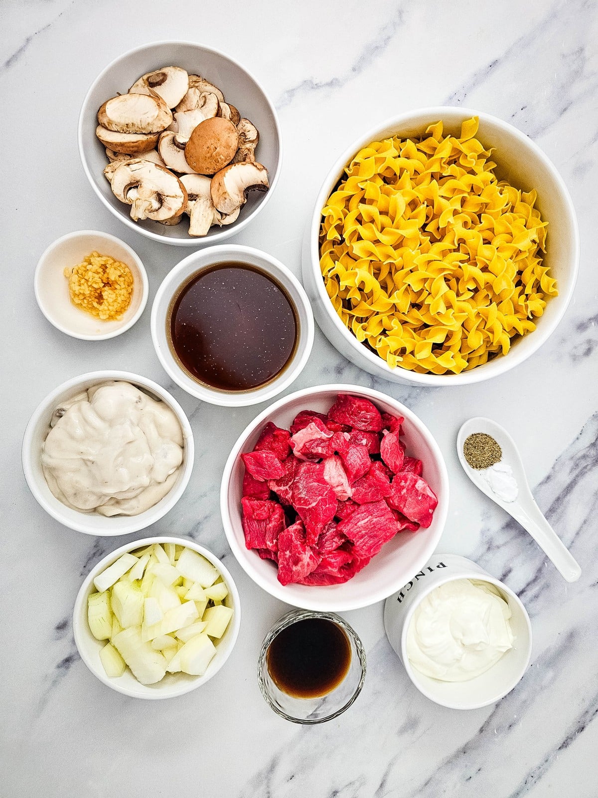 Beef stroganoff ingredients measured into bowls: stew meat, egg noodles, mushrooms, onion, sour cream, cream of mushroom soup, broth and seasonings