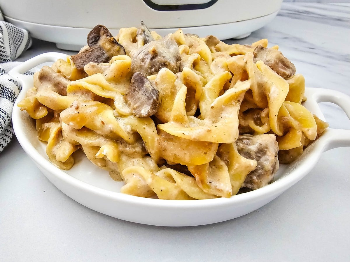 Close up of creamy beef stroganoff over egg noodles in a white dish with chunks of beef and mushroom