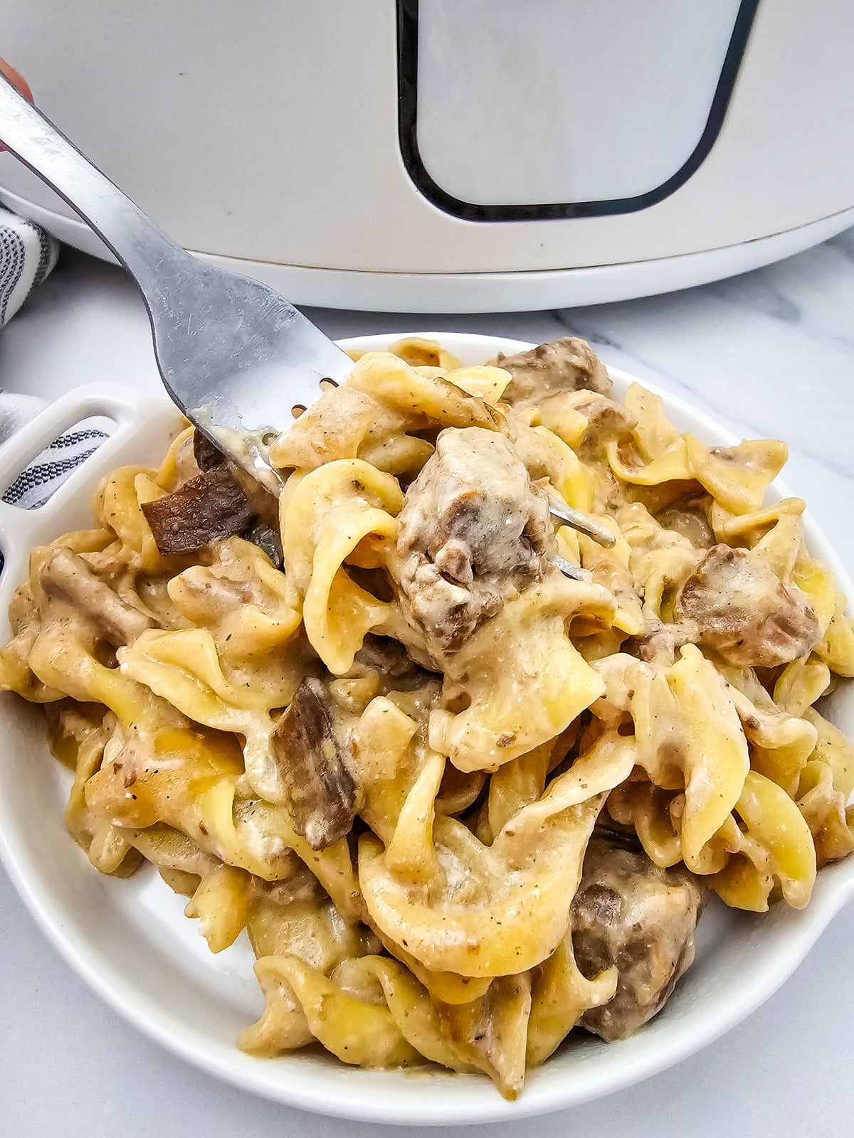 A white bowl of creamy crockpot beef stroganoff with a fork lifting egg noodles, slow cooker behind