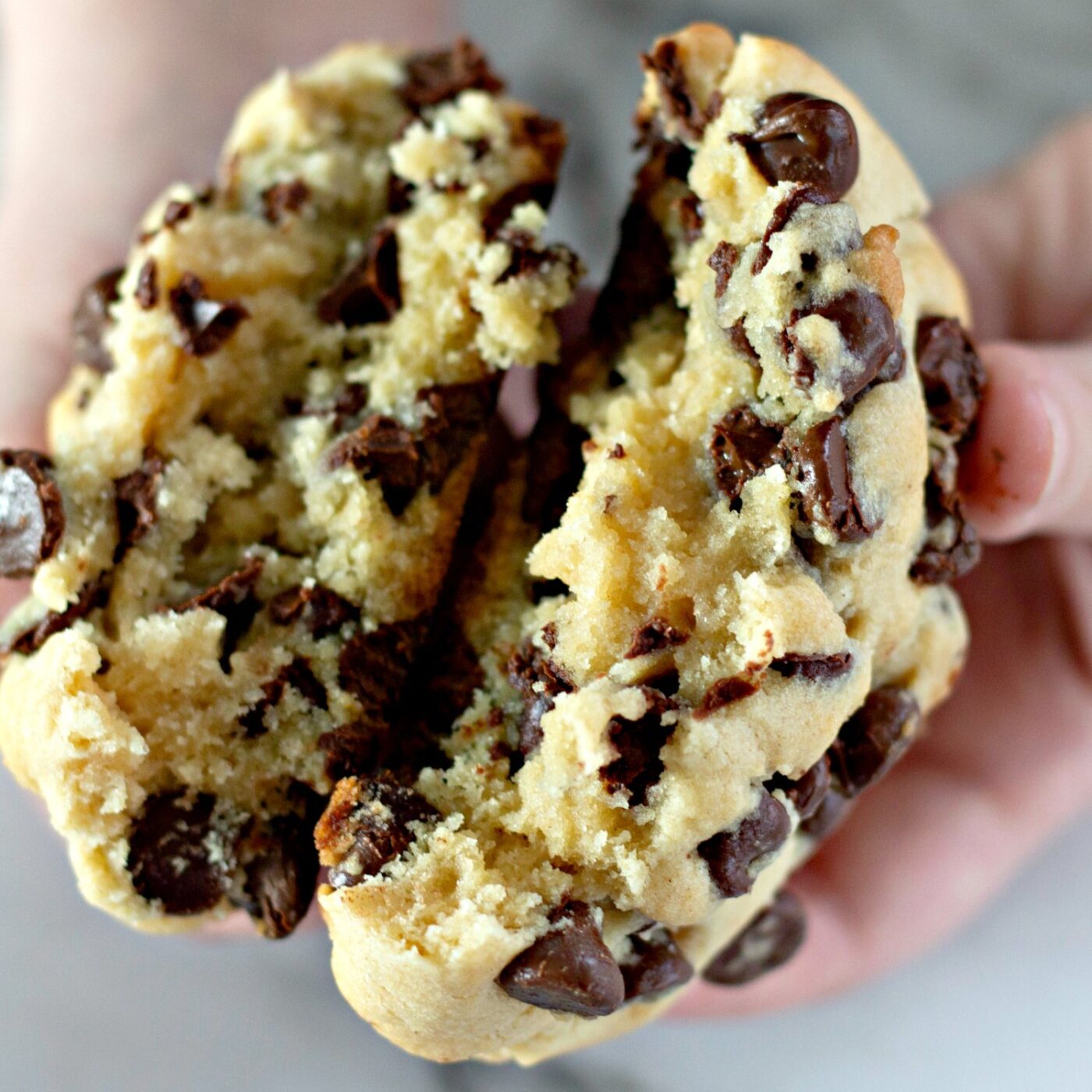Hands pulling apart a thick chocolate chip cookie to show the soft, gooey center.
