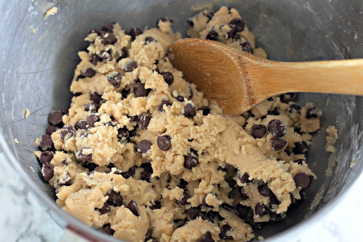 Chocolate chip cookie dough being stirred with a wooden spoon in a metal mixing bowl.