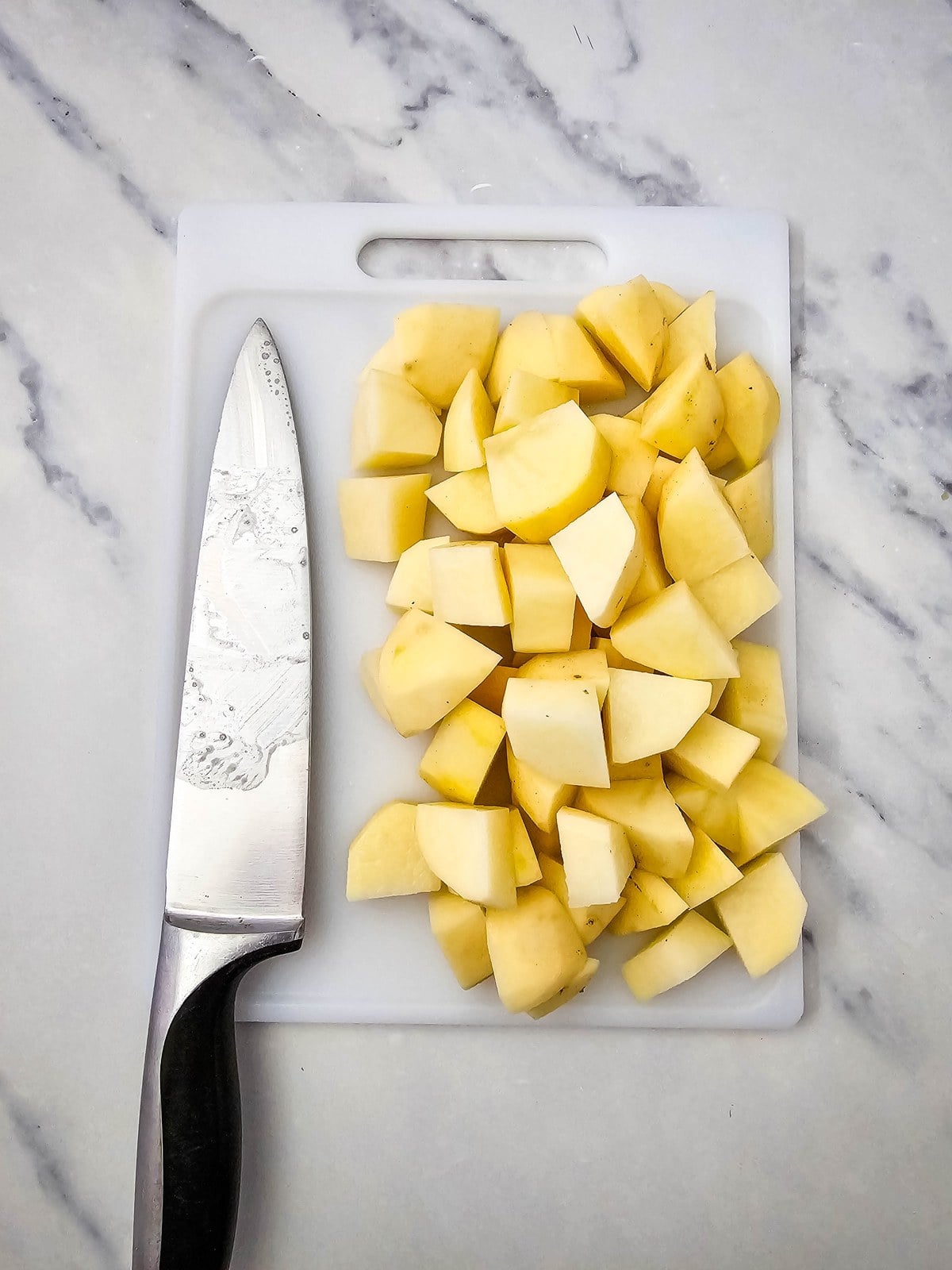 Peeled potatoes cut into one inch pieces on a white cutting board with a paring knife