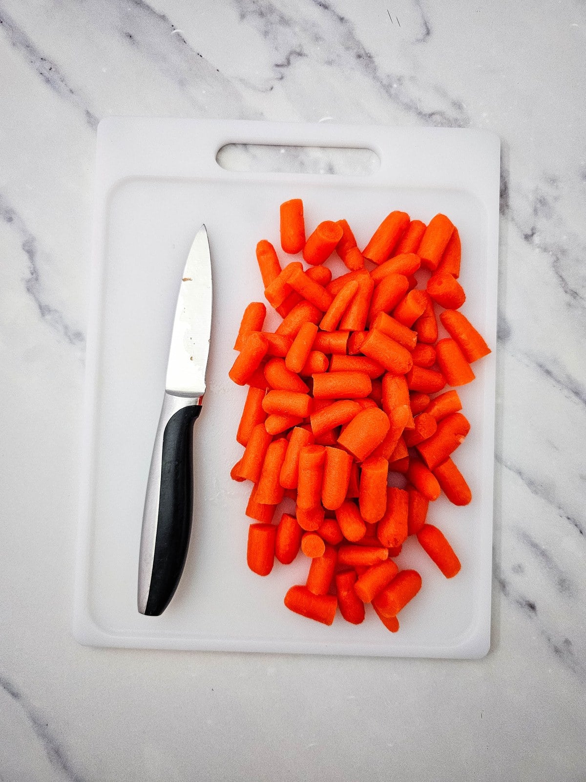 Baby carrots cut in half on a white cutting board with a paring knife