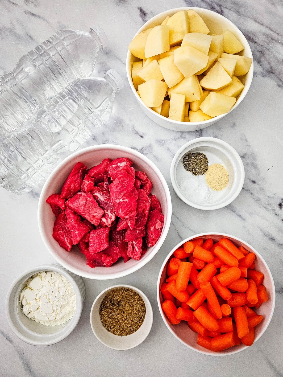 Ingredients for crockpot beef stew: water, cubed potatoes, stew meat, salt, pepper and garlic, cornstarch, beef bouillon and baby carrots in white bowls