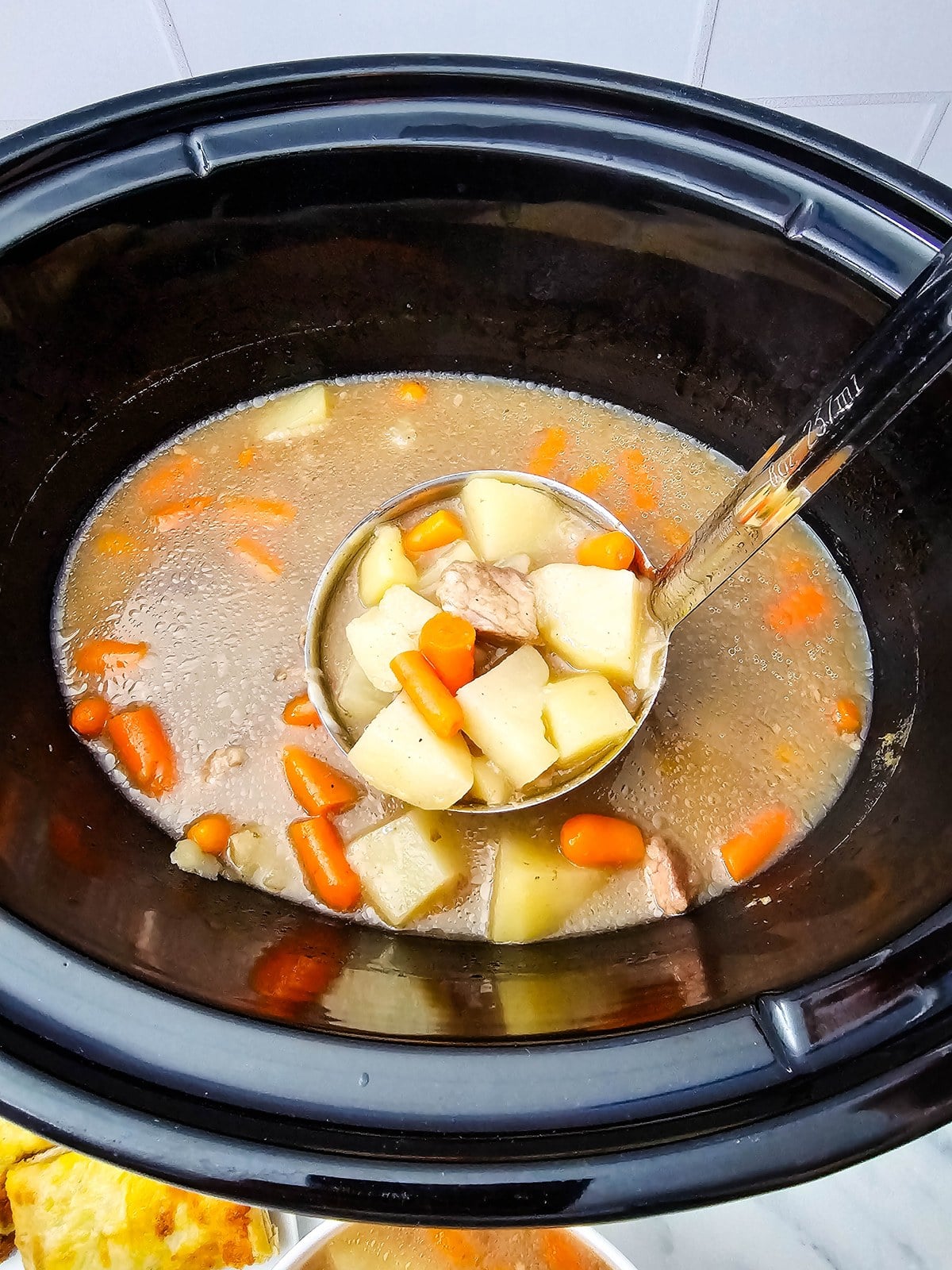 Ladle of crockpot beef stew lifted from the slow cooker full of beef, potatoes and carrots