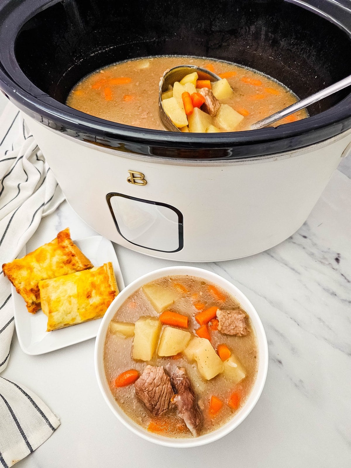Crockpot beef stew served in a white bowl beside the slow cooker with biscuits on a plate