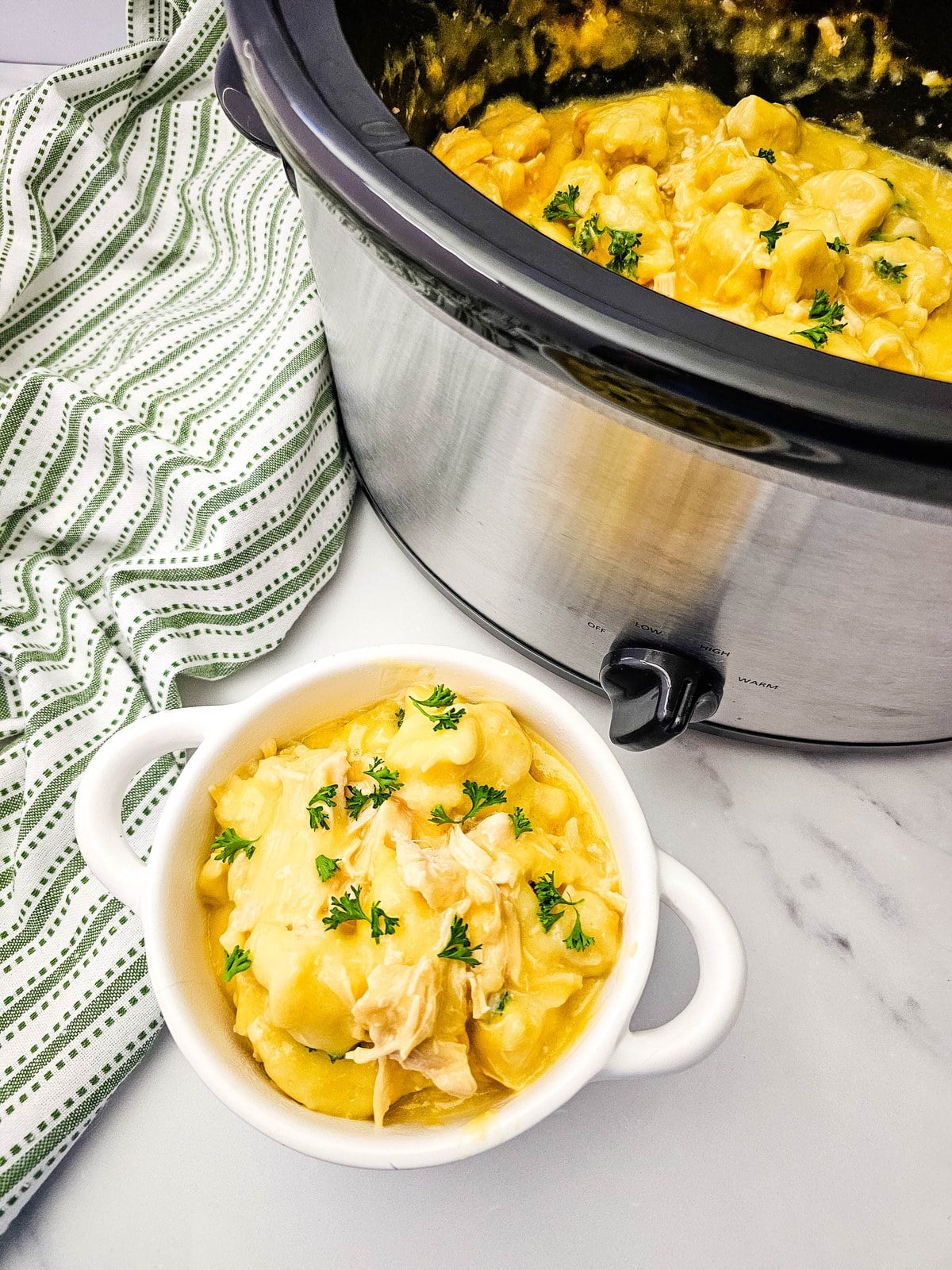 Bowl of crockpot chicken and dumplings sitting beside the slow cooker with a striped towel