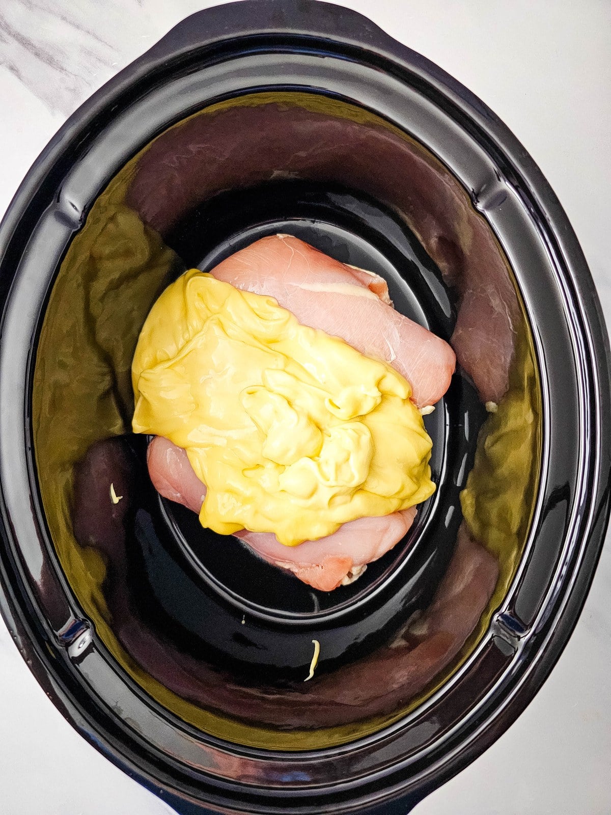 Two raw chicken breasts in a black crockpot with condensed cream of chicken soup spooned over the top
