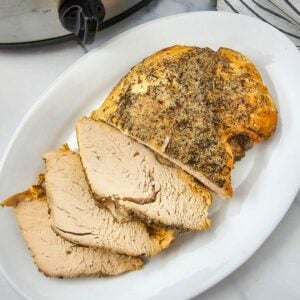 Crockpot turkey breast sliced on a white platter beside a stainless slow cooker