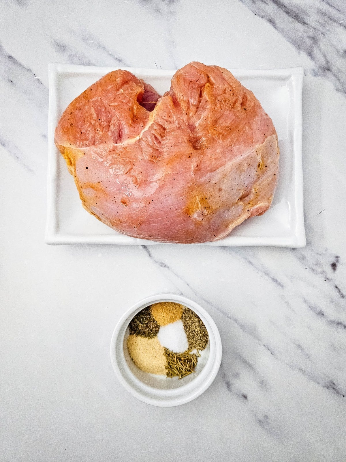 Raw boneless turkey breast on a white tray with garlic powder, onion powder, rosemary, thyme, salt and pepper in a small bowl