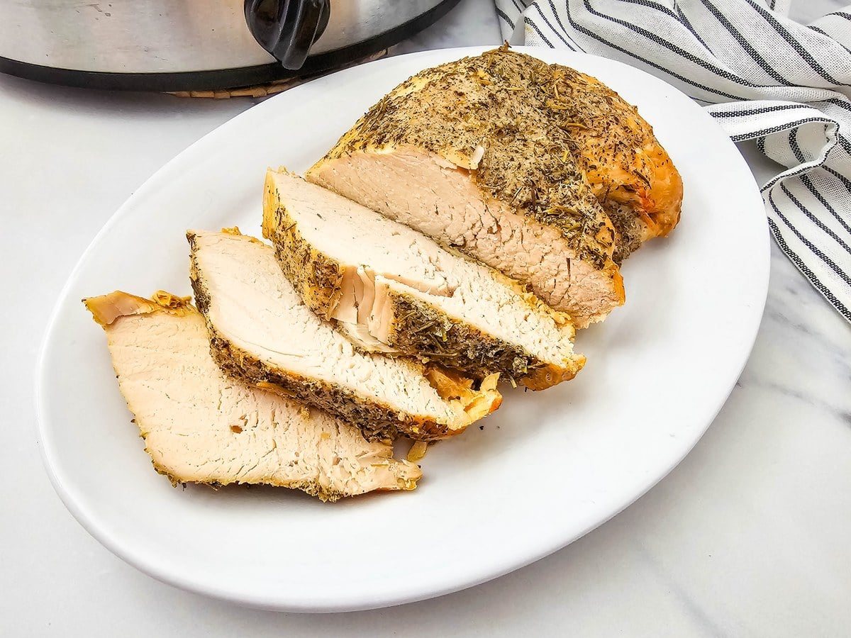 Thick slices of juicy crockpot turkey breast with an herb crust on a white platter