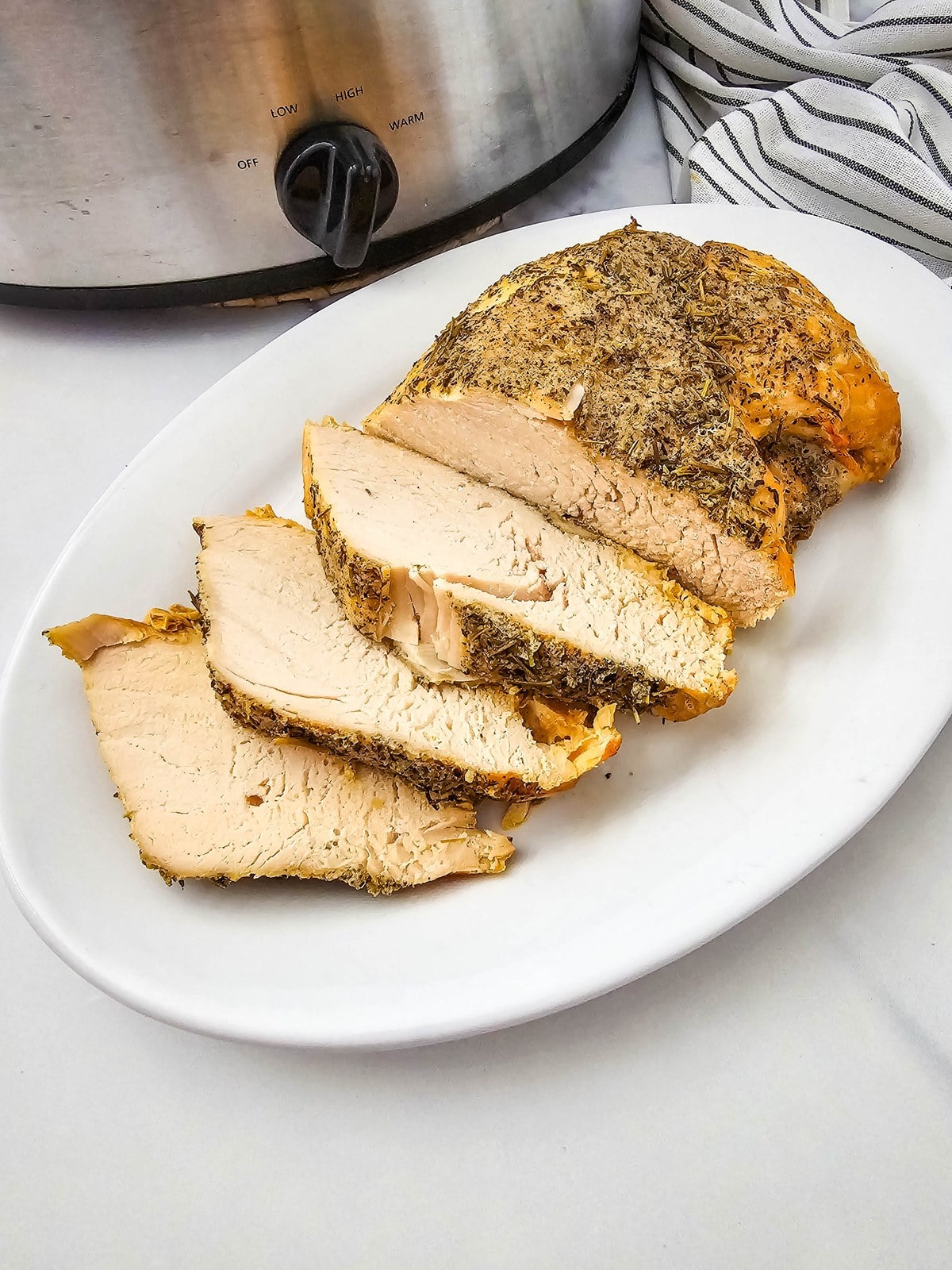 Crockpot turkey breast sliced on a white oval platter with the slow cooker behind