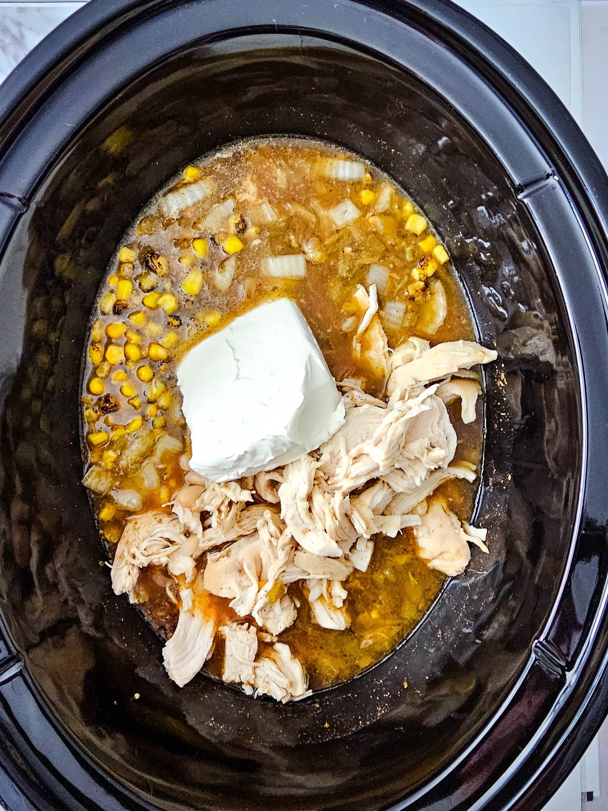 A block of cream cheese set on top of the shredded chicken in the crockpot