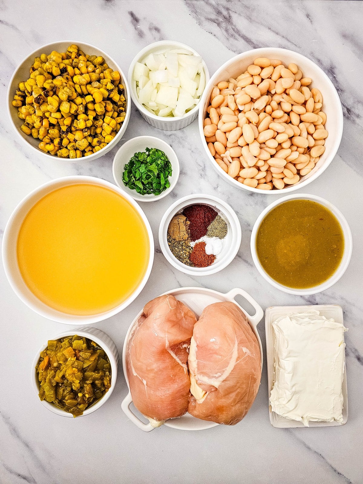 Ingredients for crockpot white chicken chili in white bowls: fire roasted corn, diced onion, white beans, cilantro, chicken broth, spices, green chili enchilada sauce, green chilis, chicken breasts and cream cheese